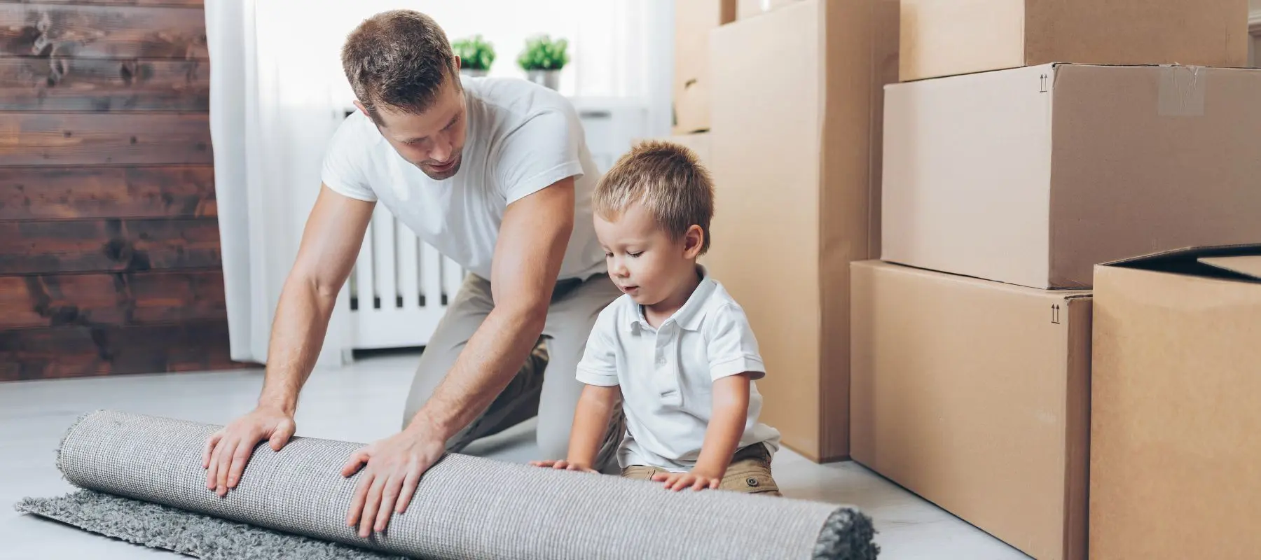 A man and a young boy roll up a rug together in a room with several cardboard boxes, suggesting they are packing or moving. Both are focused on the task. Bright natural light fills the space.