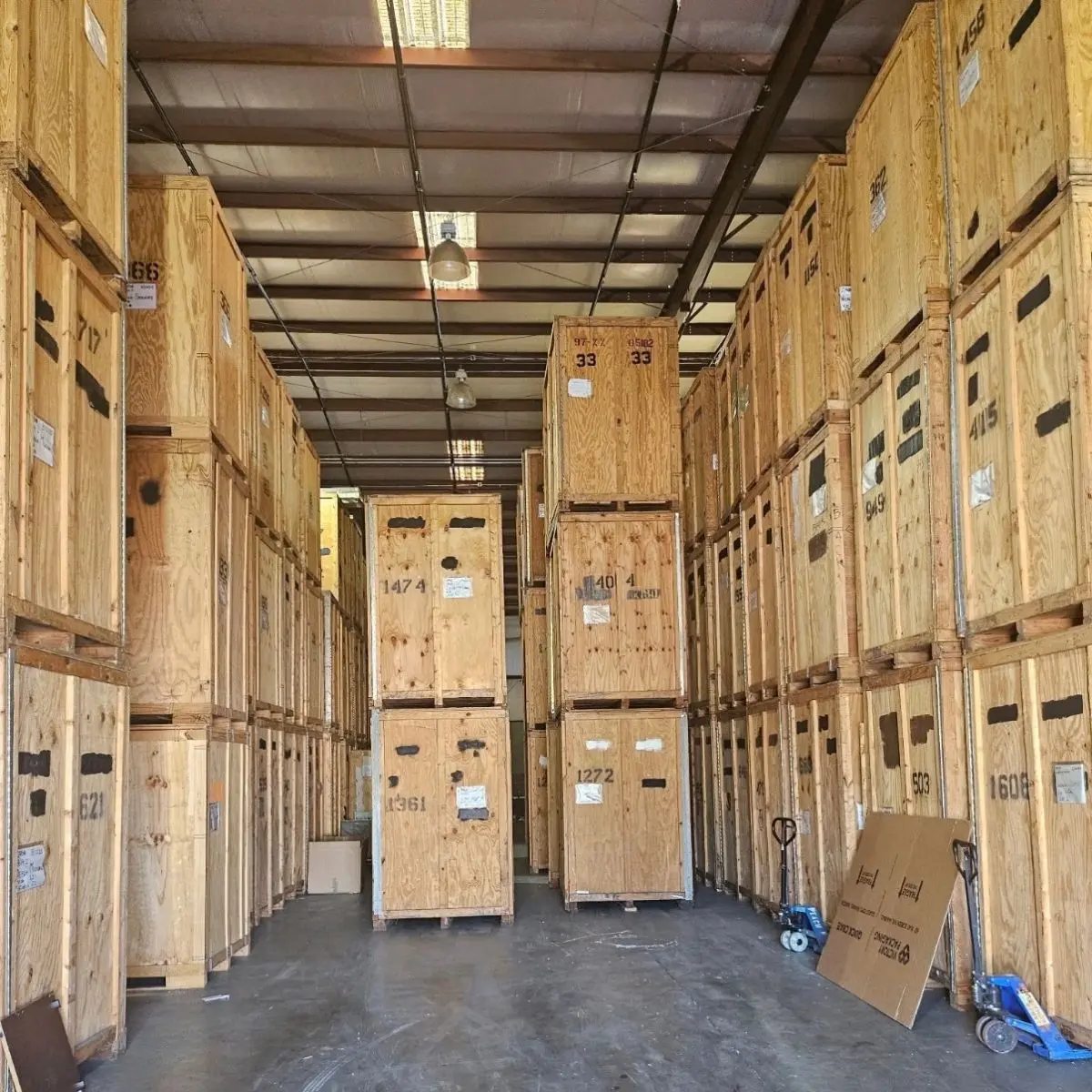 Wooden storage crates stacked high in a warehouse with a concrete floor, labels and numbers on the boxes, and several blue hand trucks and cardboard boxes near the right wall.