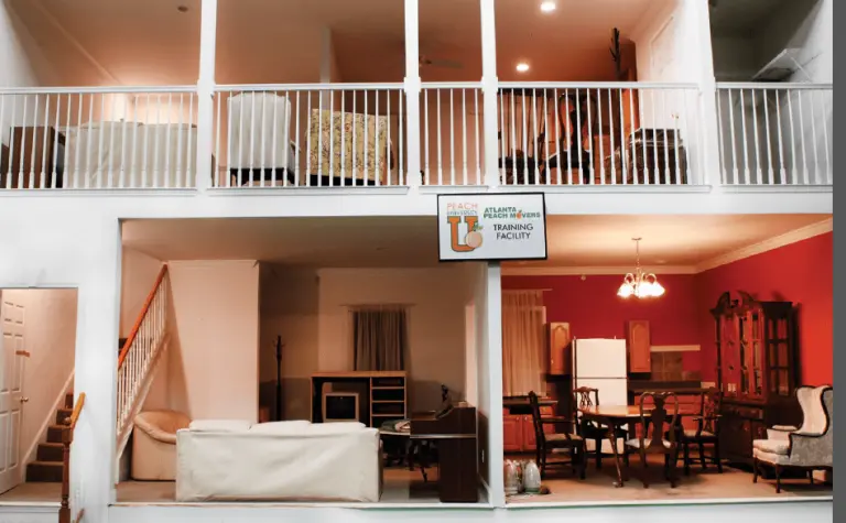 Two-story building with open walls, revealing furnished living rooms, dining area, and kitchen. A sign reads Atlanta Training Facility above a couch on the lower floor.