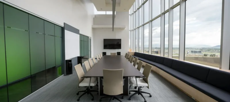 Modern conference room with a long table, chairs, large windows with city views, a black bench, a wall-mounted TV, and a whiteboard.