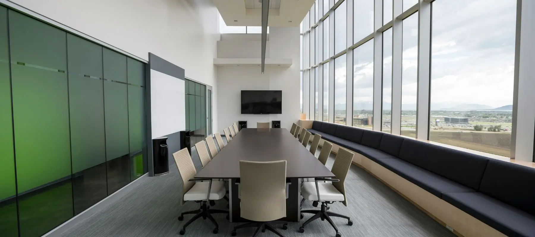 Modern conference room with a long table, chairs, large windows with city views, a black bench, a wall-mounted TV, and a whiteboard.