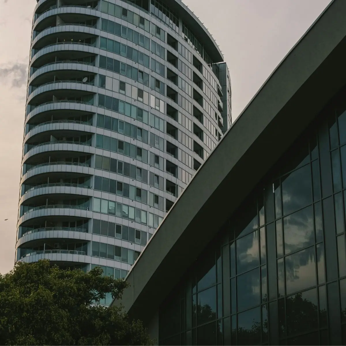 A modern high-rise building with curved balconies stands beside a glass-fronted structure, reflecting the cloudy sky and nearby trees.