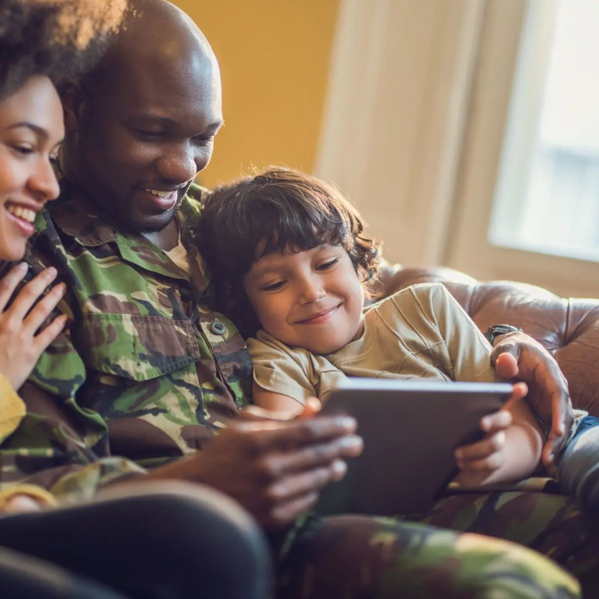 A smiling family sits on a couch together. An adult in camouflage holds a tablet, while a child and another adult sit close, all looking at the screen and appearing happy and relaxed.