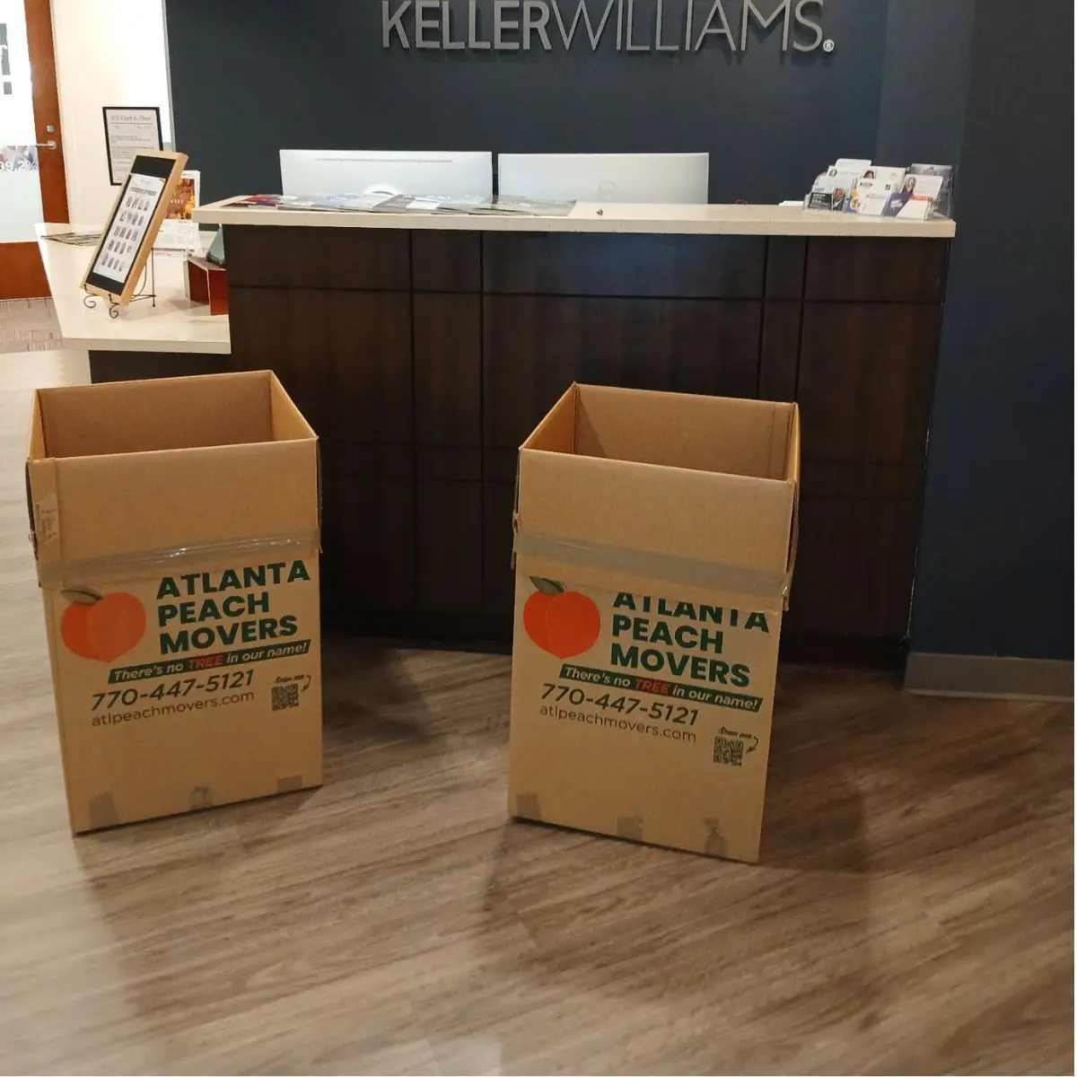 Two open moving boxes labeled Atlanta Peach Movers sit on a wood floor in front of a dark reception desk with Keller Williams signage on the wall behind.