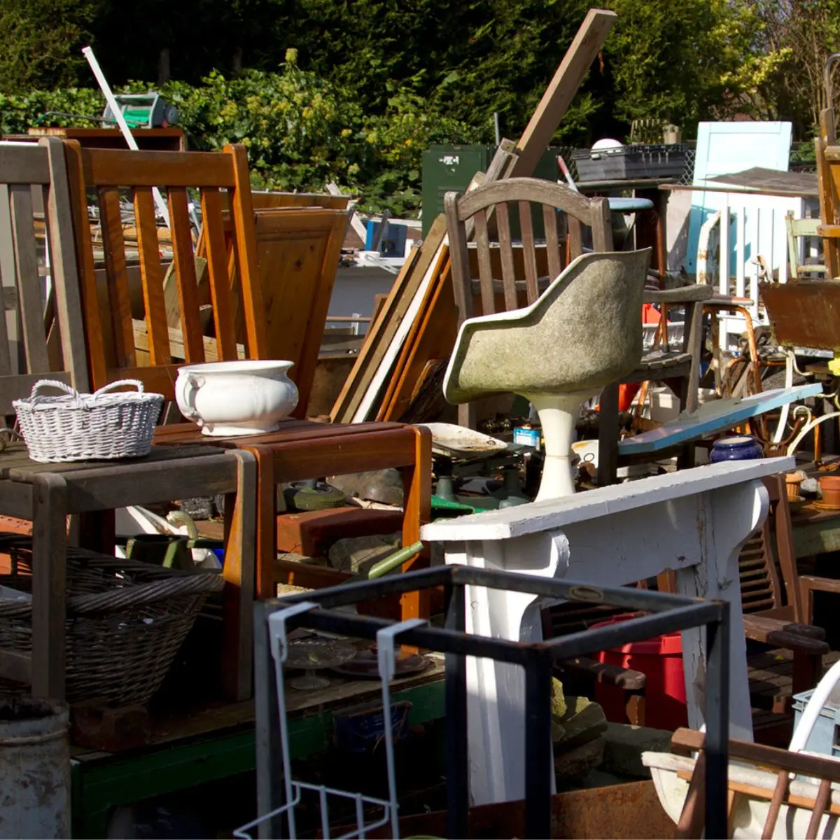 A cluttered outdoor area filled with old chairs, tables, baskets, and various discarded household items in disarray, likely at a junkyard or salvage yard. Greenery and trees are visible in the background.