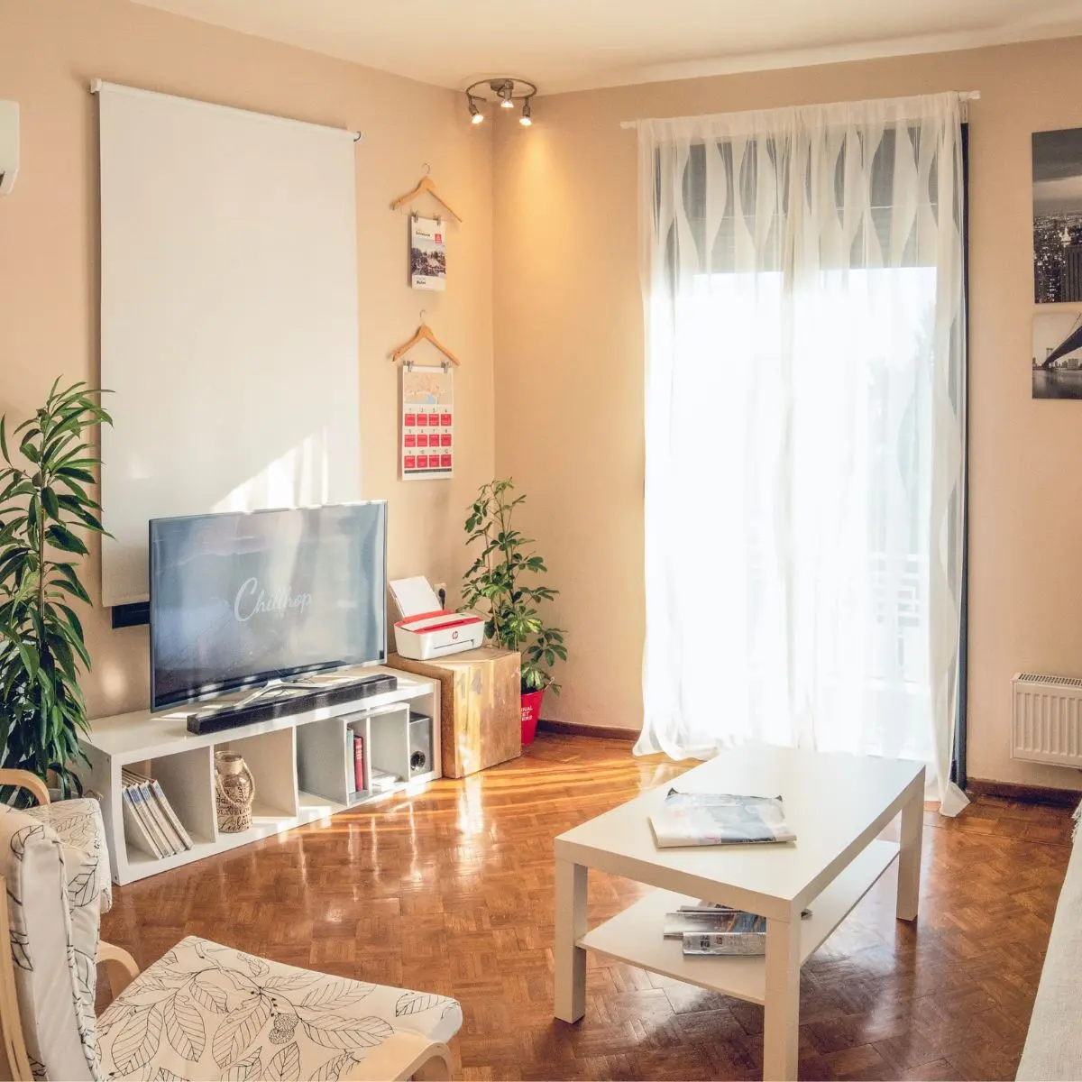 Bright living room with a TV, white furniture, potted plants, a large window with sheer curtains, wooden floors, and wall decor. Sunlight streams in, creating a cozy, inviting atmosphere.