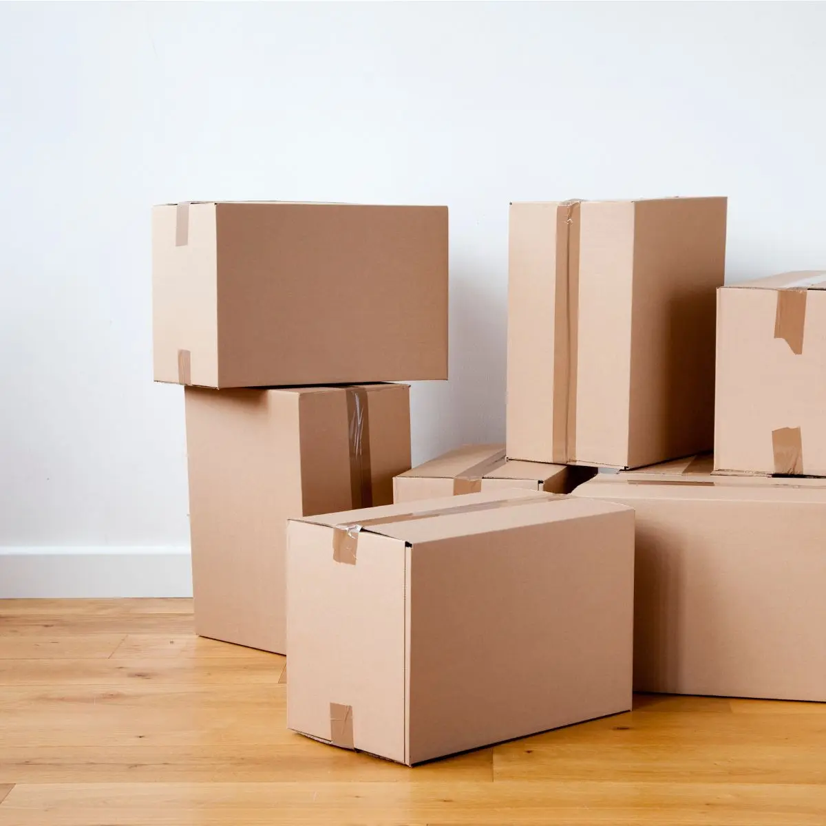 Six closed cardboard boxes of various sizes are stacked on a hardwood floor next to a white wall, suggesting moving or packing.