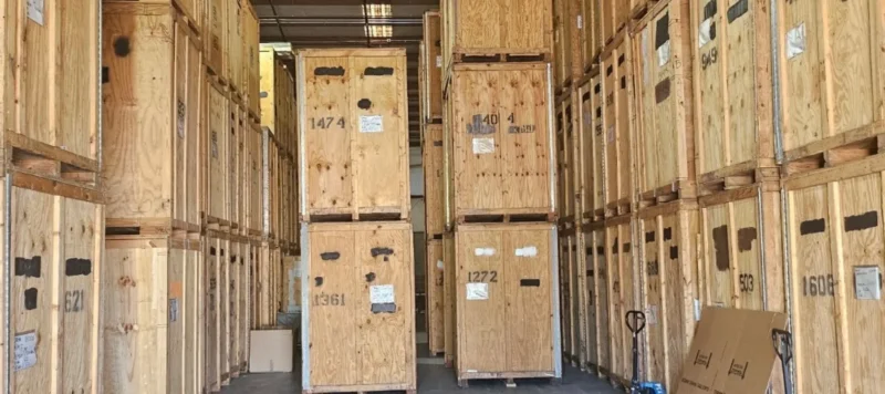 Tall stacks of large wooden crates line both sides of a warehouse aisle, with labels and numbers on each crate. A pallet jack and cardboard boxes are visible near the bottom right corner.