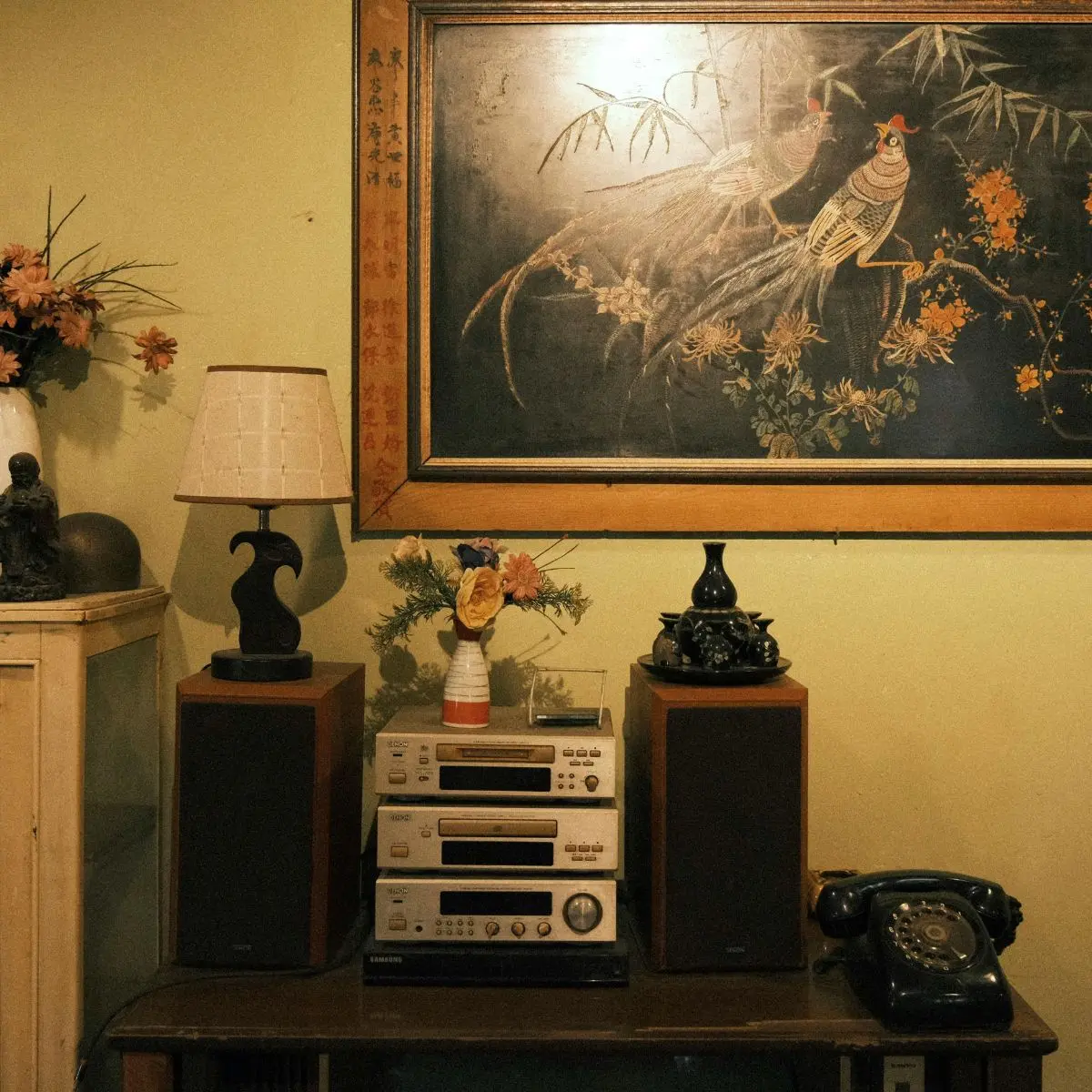 A vintage living room setup with a stereo system, old rotary phone, and table lamp. Above them hangs an ornate painting of birds on a branch. Flowers decorate the area, adding a cozy touch.